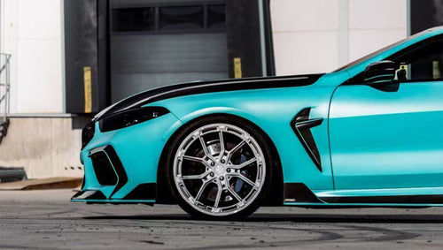 Turquoise BMW F92 M8 featuring a REVOZPORT carbon fiber body kit with front lip, side skirts, black hood accents, side vents, and silver forged wheels parked outside an industrial loading dock.