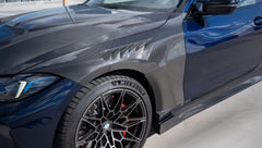 Black BMW G82 M4 featuring a REVOZPORT carbon fiber body kit with front lip, side skirts, vented fenders, and aggressive aero styling parked on a rooftop with city buildings in the background.
