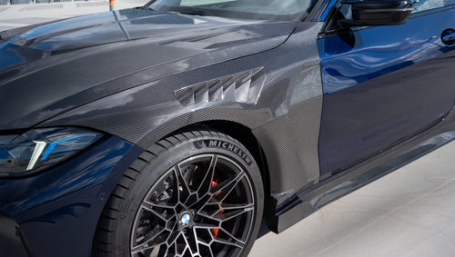 Black BMW G82 M4 featuring a REVOZPORT carbon fiber body kit with front lip, side skirts, vented fenders, and aggressive aero styling parked on a rooftop with city buildings in the background.