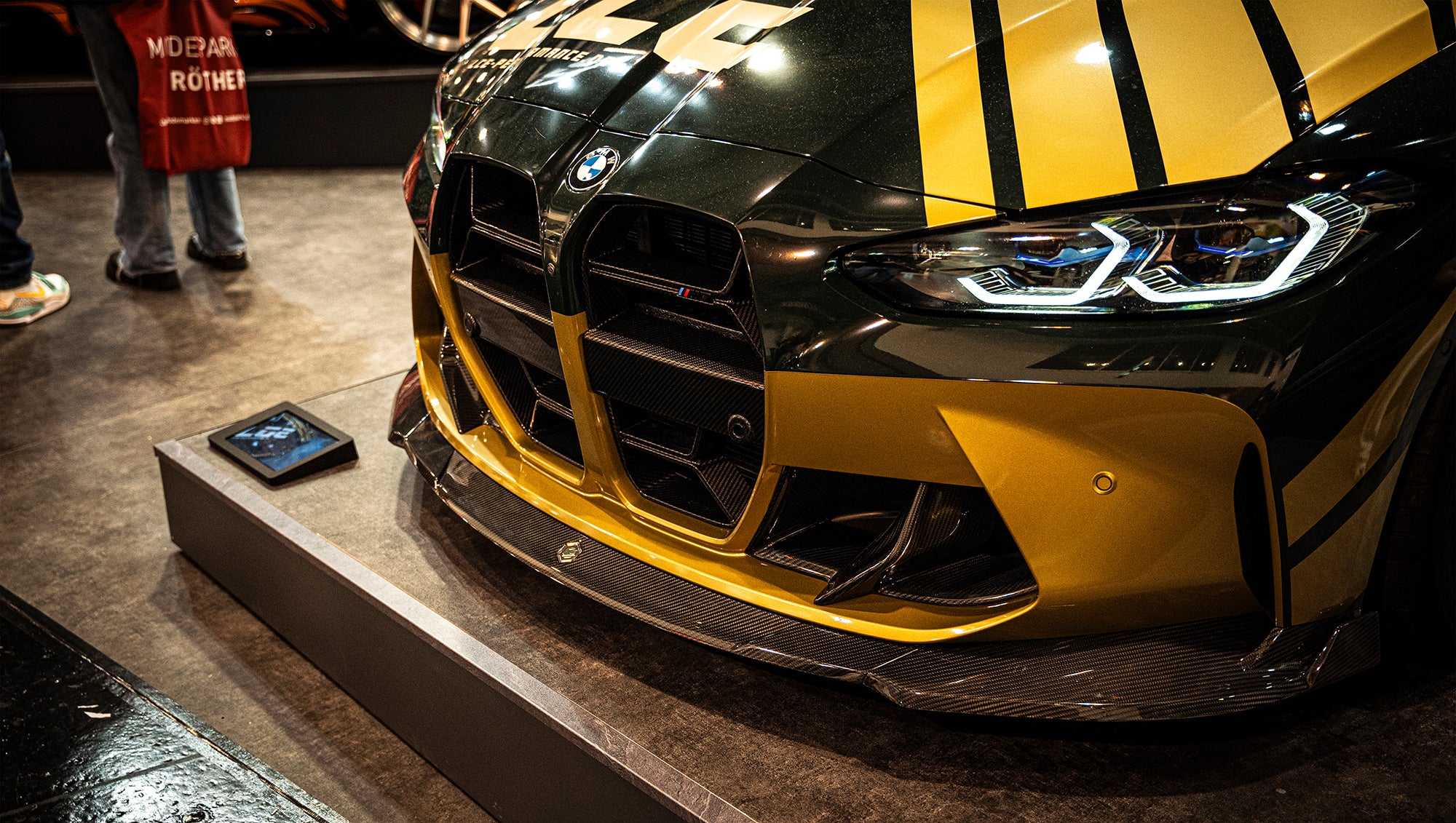 BMW G80 M3 race build with REVOZPORT carbon fiber aero kit, gold wheels, and black-yellow motorsport livery displayed at an indoor automotive exhibition.