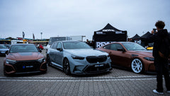 Gray BMW G90 M5 Touring featuring a REVOZPORT carbon fiber front lip, aggressive front aero styling, and black grille displayed at a car event.