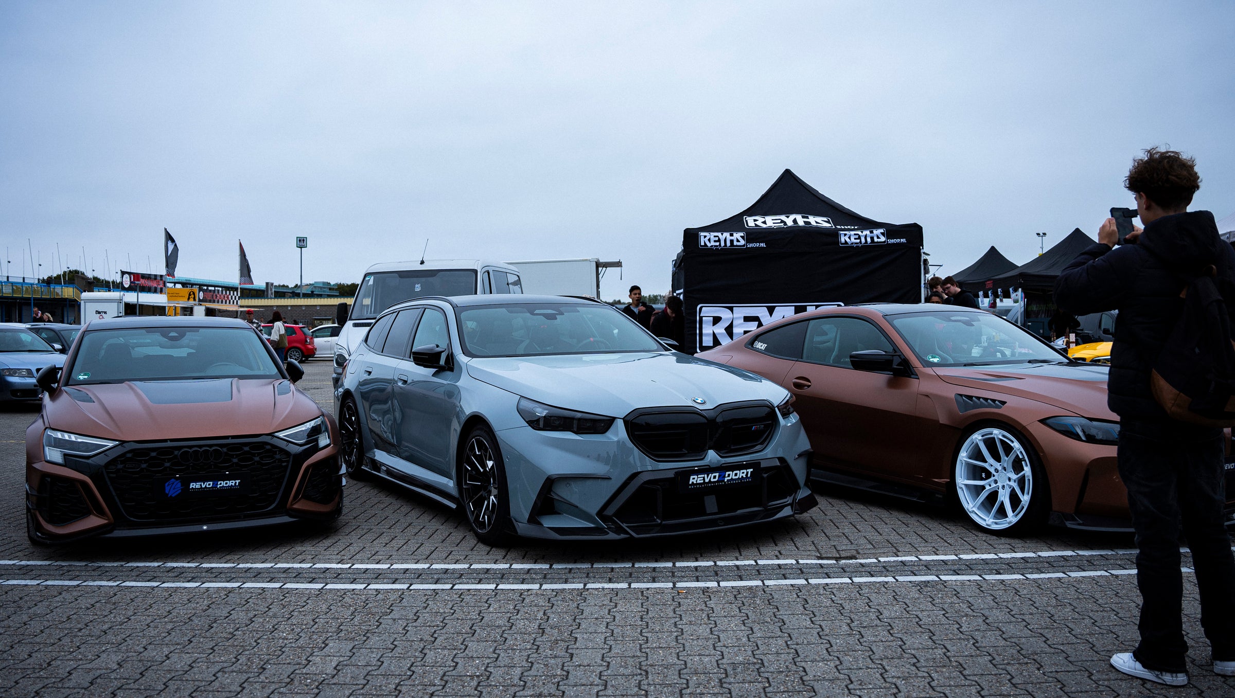 Gray BMW G90 M5 Touring featuring a REVOZPORT carbon fiber front lip, aggressive front aero styling, and black grille displayed at a car event.