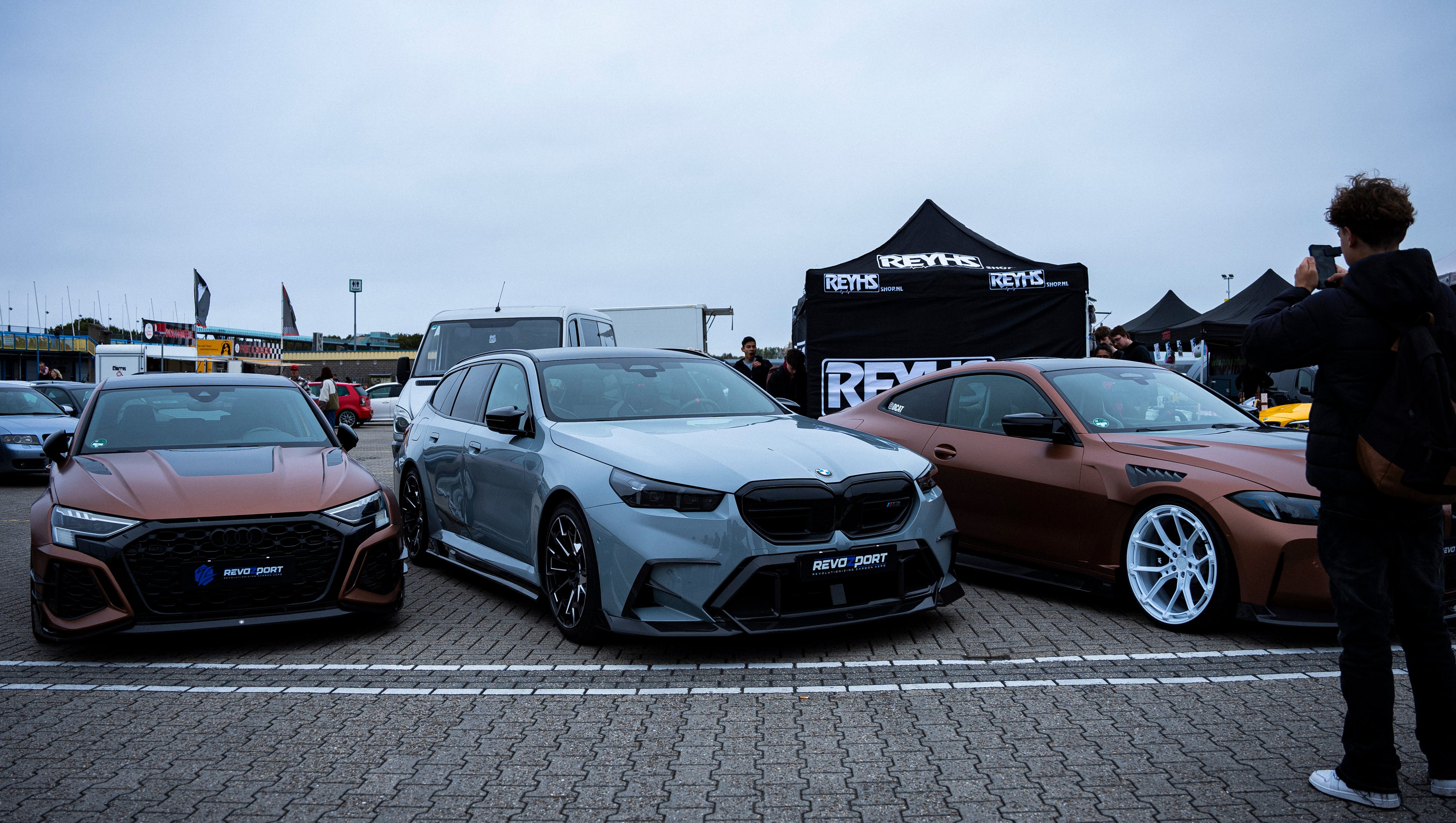 Gray BMW G90 M5 Touring featuring a REVOZPORT carbon fiber front lip, aggressive front aero styling, and black grille displayed at a car event.