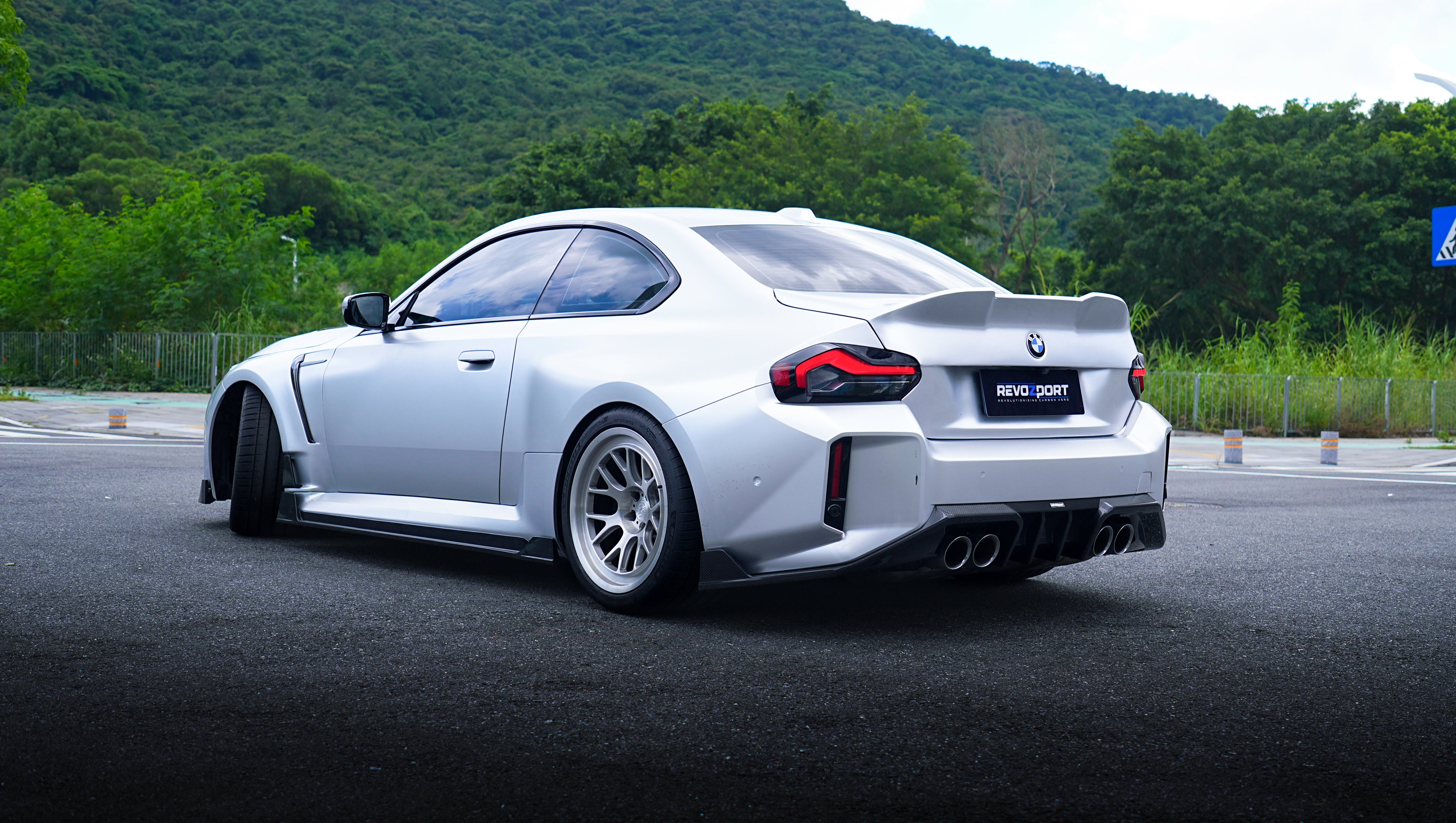 Front view of a silver BMW G87 M2 featuring a REVOZPORT carbon fiber front lip, aggressive front bumper aero styling, and vented hood parked on an urban street.