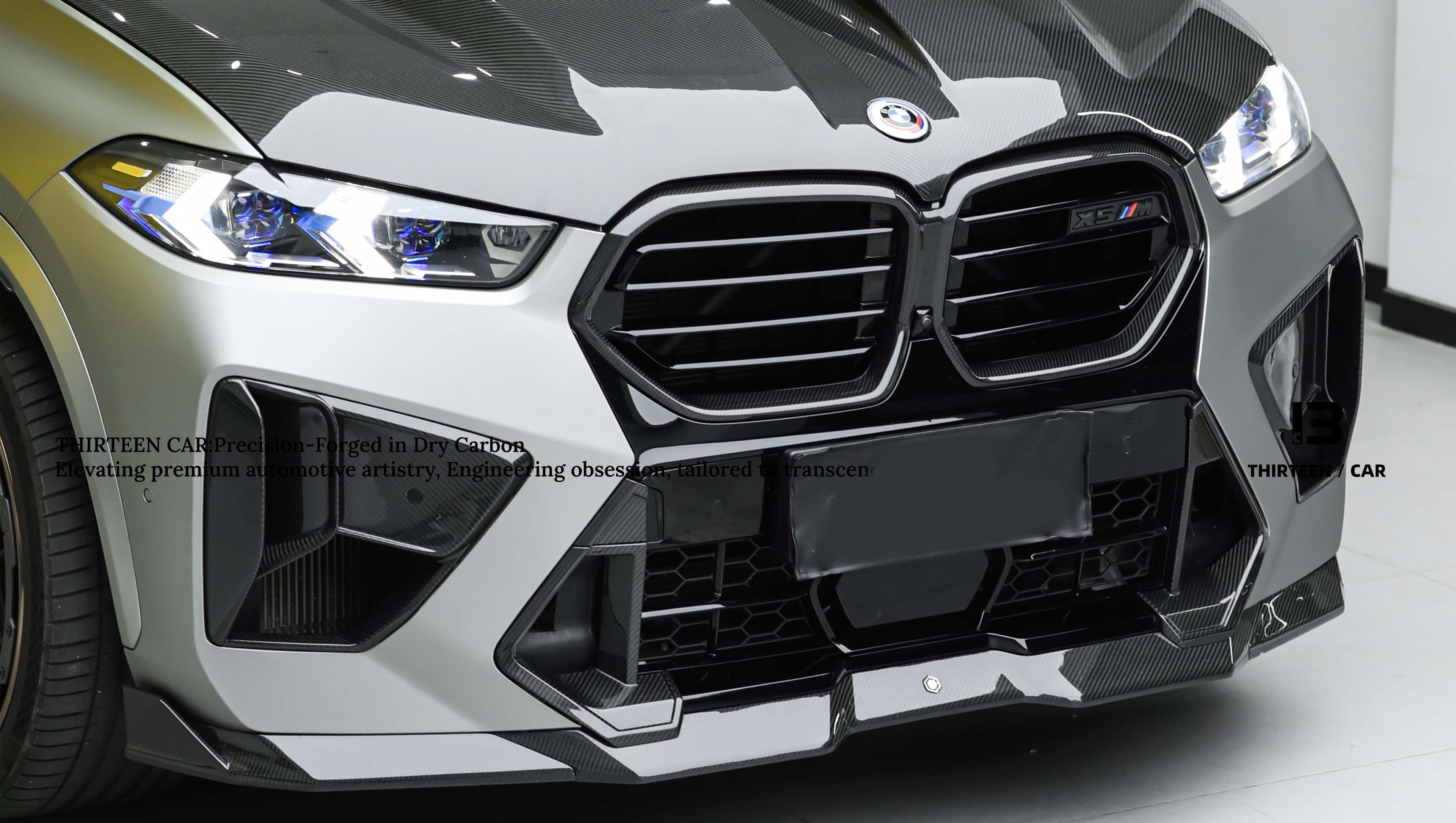Gray BMW X5M featuring a REVOZPORT carbon fiber body kit with aggressive front lip, enlarged air intakes, side skirts, and sporty aero styling displayed inside a performance workshop.