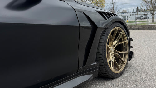 Close-up of a matte black BMW G82 M4 featuring REVOZPORT carbon fiber vented fenders, carbon side skirts, bronze forged wheels, and red performance brakes.