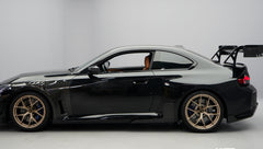 Front close-up of a black BMW G87 M2 featuring an aggressive carbon fiber front bumper, deep front splitter, large air intakes, and gloss carbon fiber aero details in a studio setting.