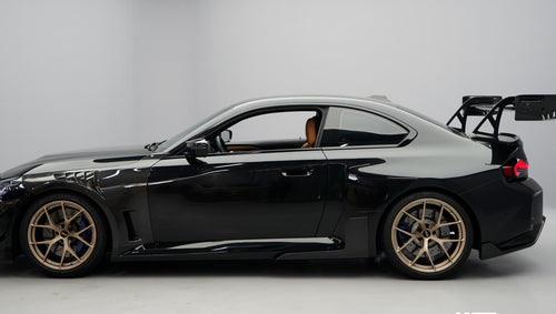 Front close-up of a black BMW G87 M2 featuring an aggressive carbon fiber front bumper, deep front splitter, large air intakes, and gloss carbon fiber aero details in a studio setting.