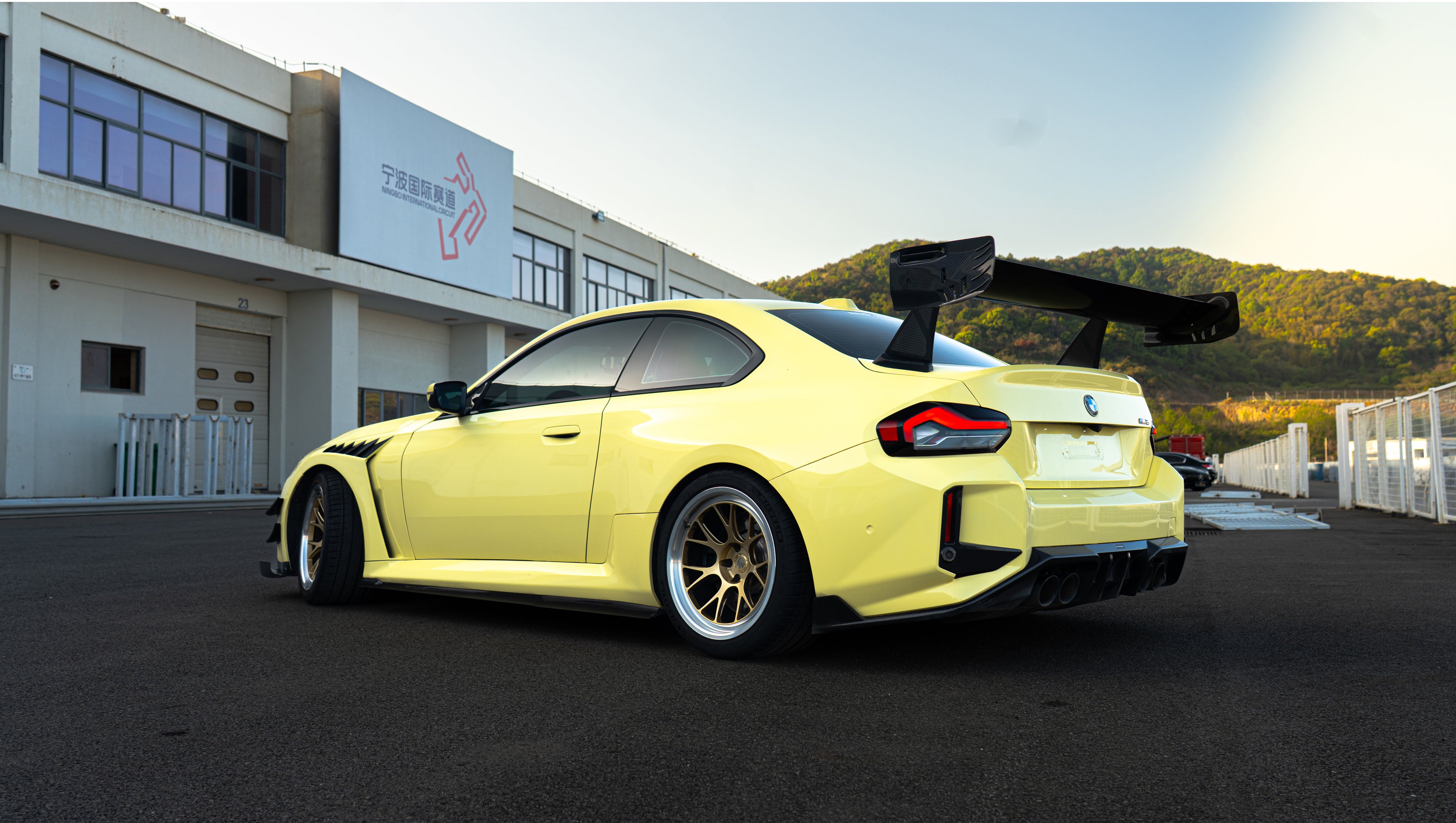 Pastel yellow BMW G82 M4 with carbon fiber front splitter, hood vents, side canards, bronze wheels, and large rear wing parked at a race track facility during golden hour.