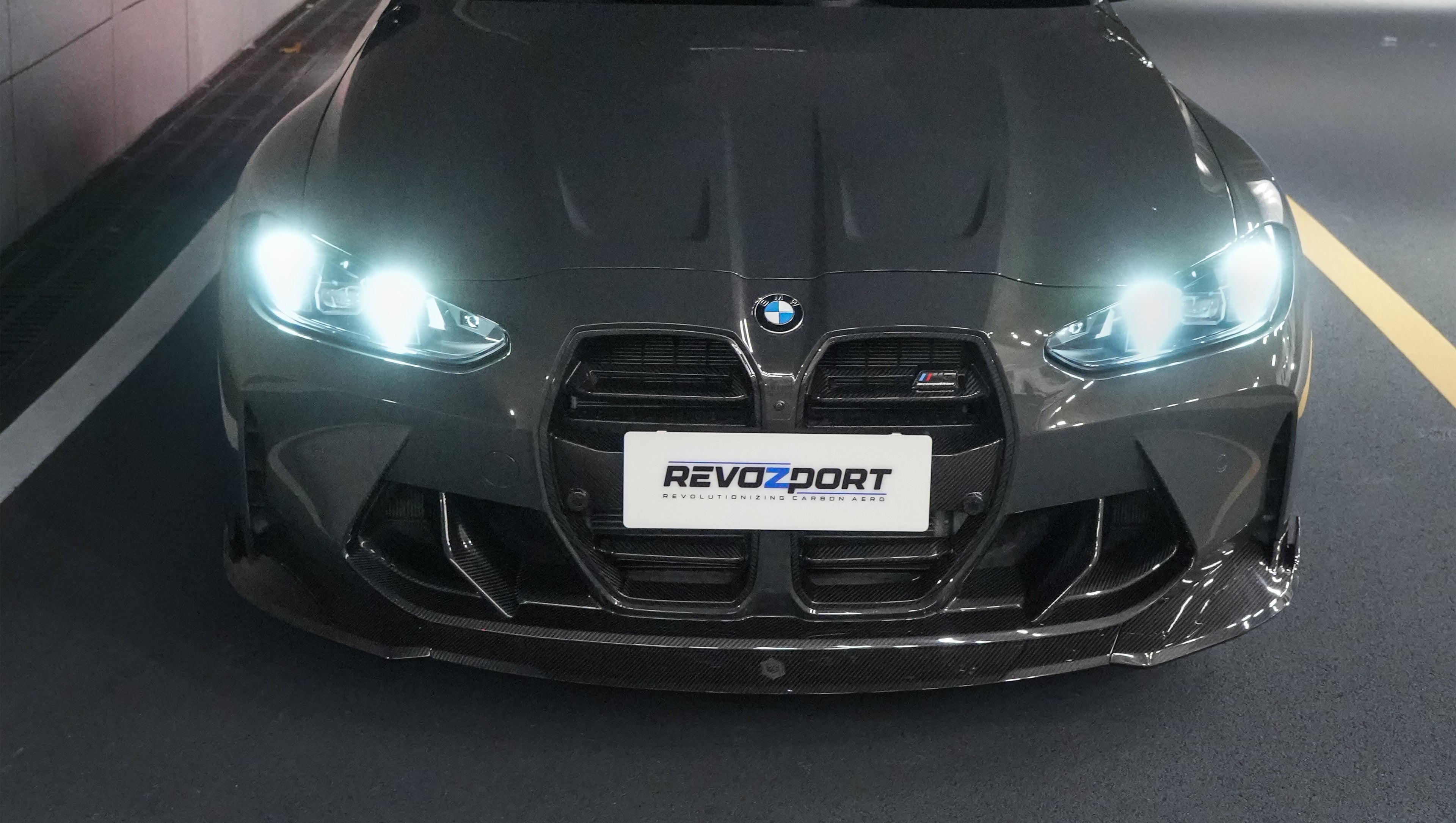 Gray BMW G80 M3 featuring a REVOZPORT carbon fiber front lip, side skirts, bronze wheels, and M The M3 side graphics driving through a tunnel at night.