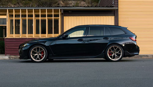 Black BMW G81 M3 Touring featuring a REVOZPORT carbon fiber body kit with vented fenders, side skirts, bronze wheels, and red performance brakes parked beside a stone wall.