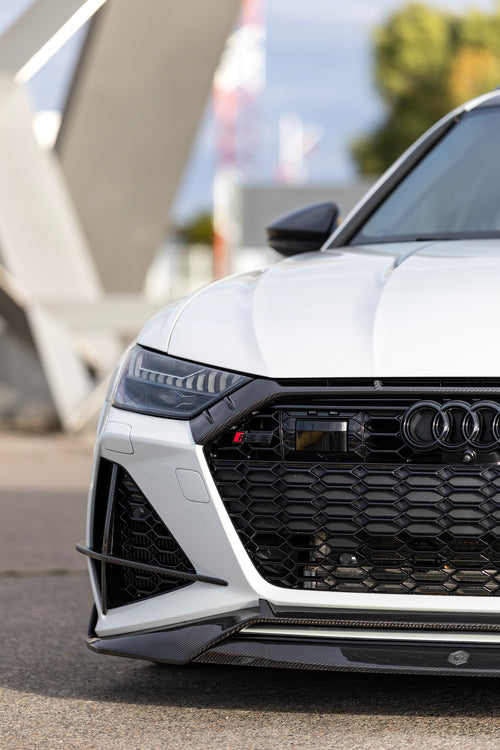 White Audi RS6 Avant featuring a REVOZPORT carbon fiber body kit with front lip, side skirts, and aggressive aero styling parked in front of a historic stone mansion during autumn.