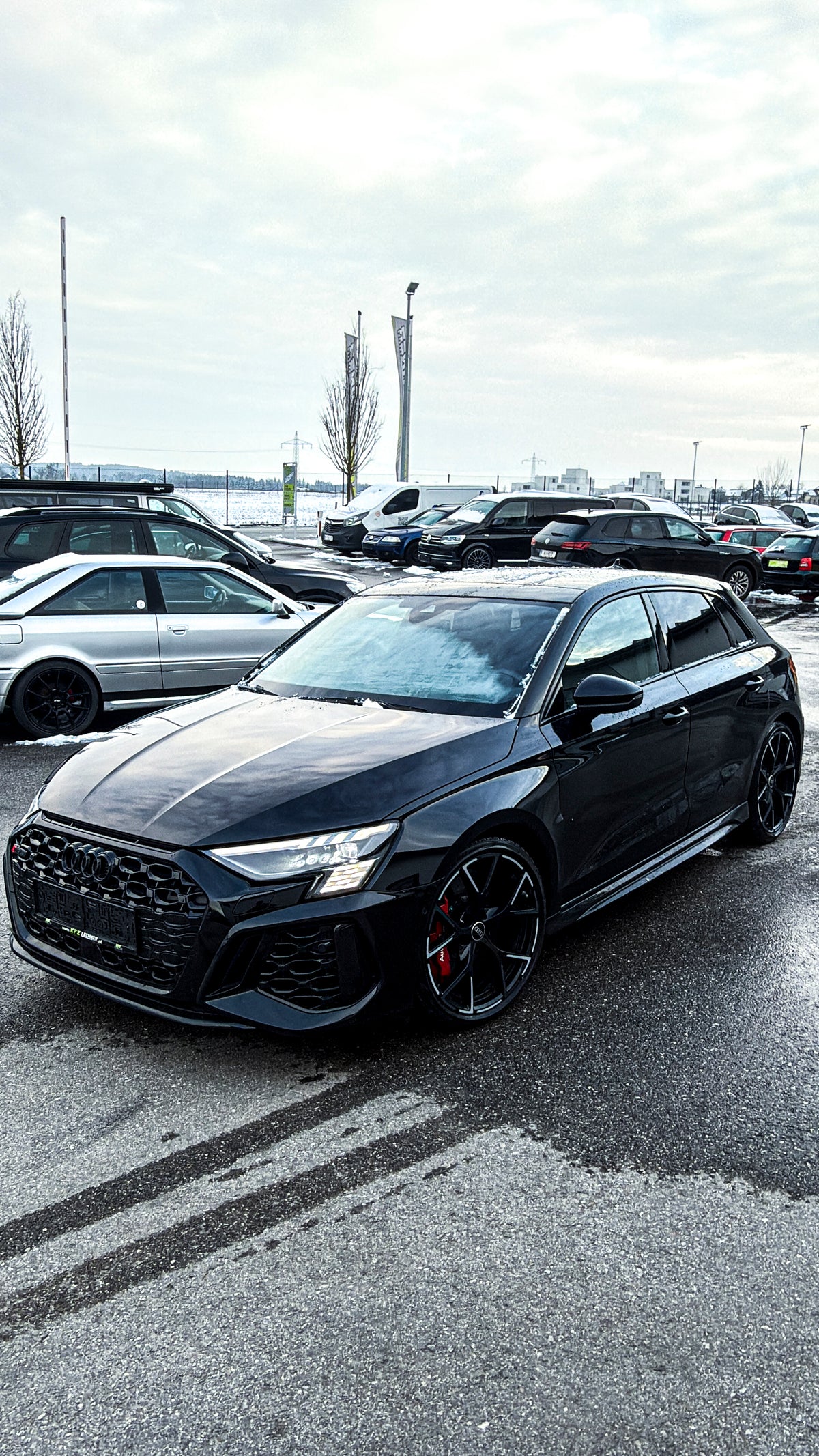 Black Audi RS3 Sportback featuring a REVOZPORT carbon fiber body kit with front lip and side skirts parked on a wet outdoor lot.