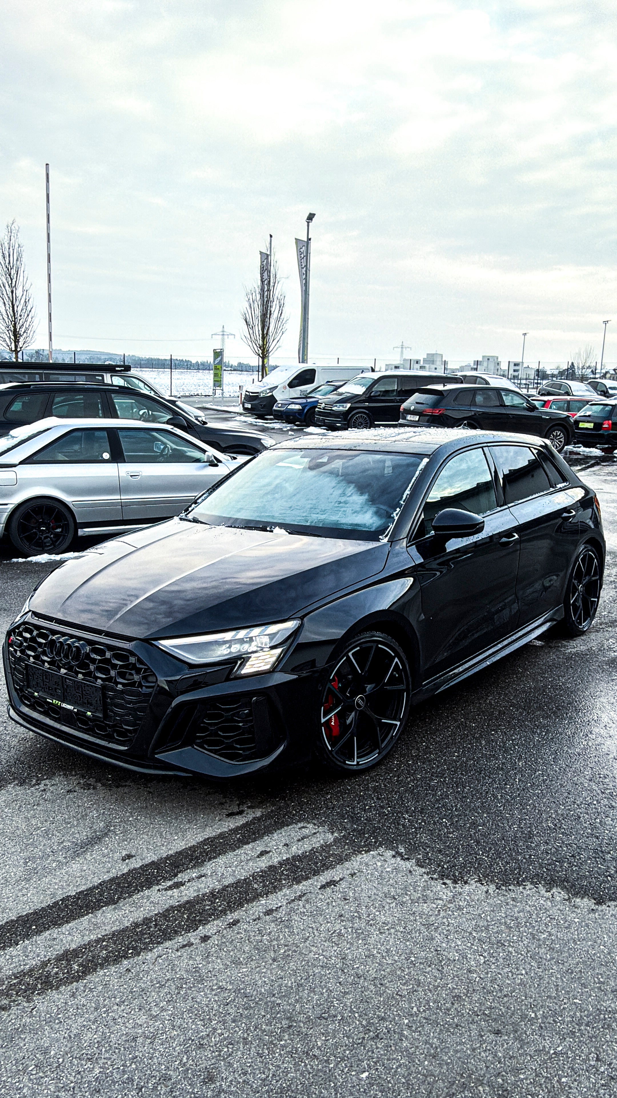 Black Audi RS3 Sportback featuring a REVOZPORT carbon fiber body kit with front lip and side skirts parked on a wet outdoor lot.