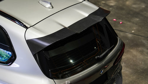 Gray BMW X5M featuring a REVOZPORT carbon fiber body kit with aggressive front lip, enlarged air intakes, side skirts, and sporty aero styling displayed inside a performance workshop.