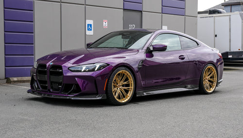 Purple BMW G82 M4 featuring a REVOZPORT carbon fiber body kit with front lip, side skirts, vented fenders, and gold wheels parked outside a modern industrial building.