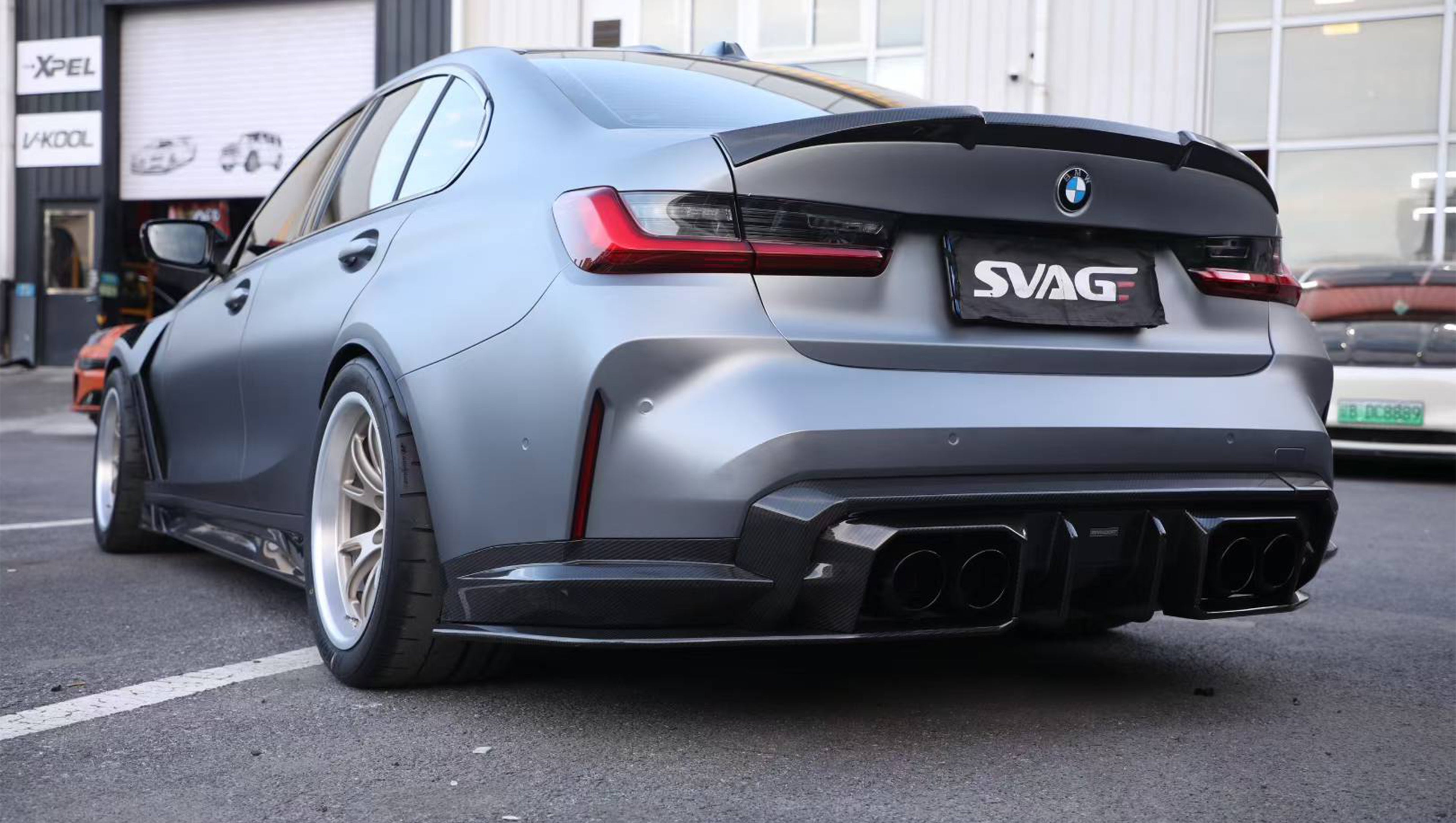 Close-up of a gray BMW G80 M3 featuring a REVOZPORT carbon fiber front lip, vented fenders, side skirts, and aggressive front aero styling in an outdoor parking lot.