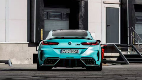Turquoise BMW F92 M8 featuring a REVOZPORT carbon fiber body kit with front lip, side skirts, black hood accents, side vents, and silver forged wheels parked outside an industrial loading dock.