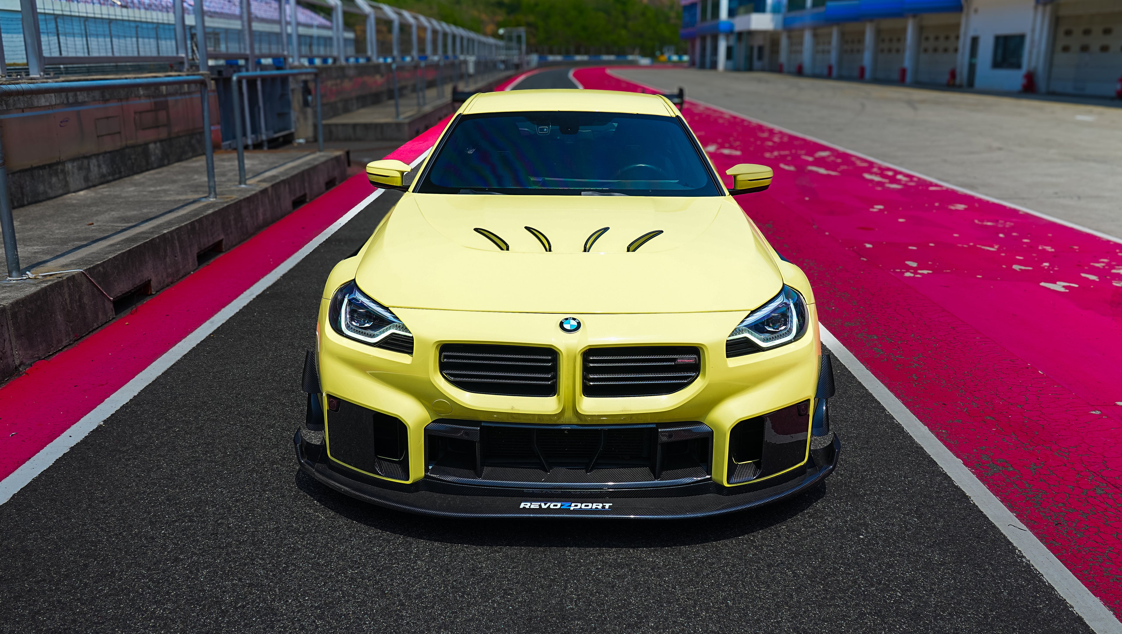 Yellow BMW G82 M4 with aggressive carbon fiber front splitter, vented hood, side canards, and bronze wheels parked on a race track pit lane.