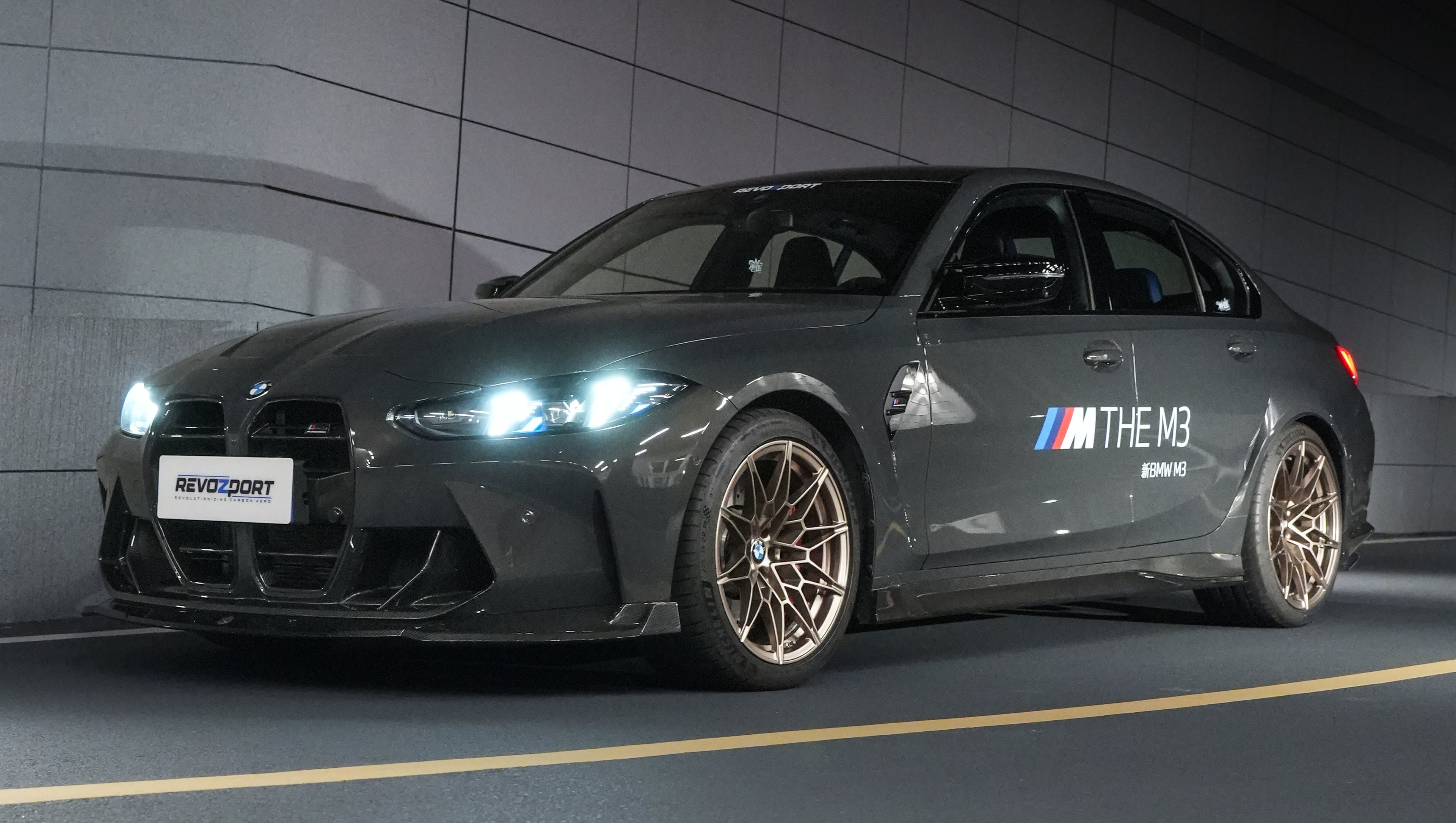 Gray BMW G80 M3 featuring a REVOZPORT carbon fiber front lip, side skirts, bronze wheels, and M The M3 side graphics driving through a tunnel at night.