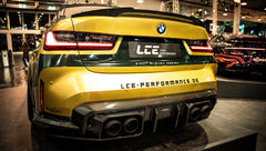 BMW G80 M3 race build with REVOZPORT carbon fiber aero kit, gold wheels, and black-yellow motorsport livery displayed at an indoor automotive exhibition.