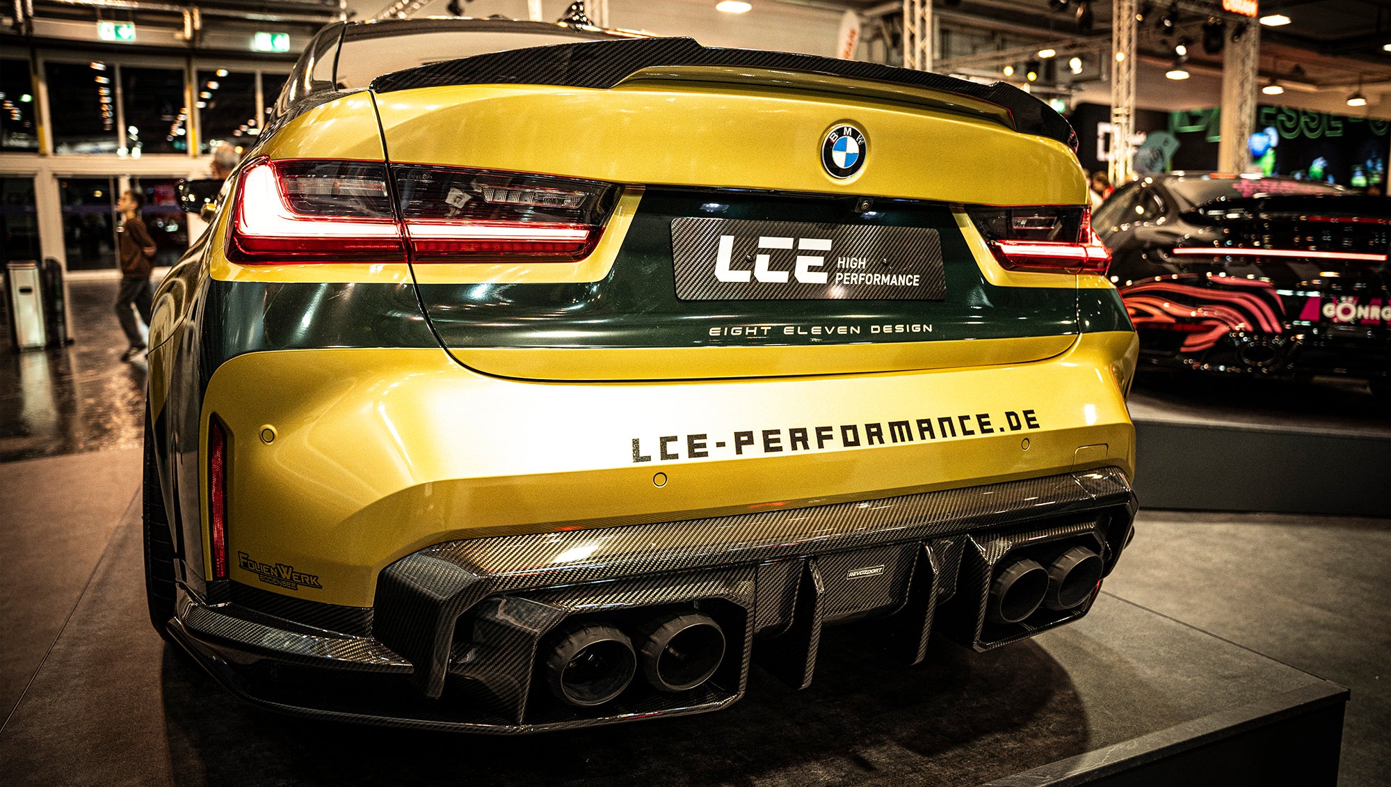 BMW G80 M3 race build with REVOZPORT carbon fiber aero kit, gold wheels, and black-yellow motorsport livery displayed at an indoor automotive exhibition.