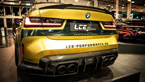 BMW G80 M3 race build with REVOZPORT carbon fiber aero kit, gold wheels, and black-yellow motorsport livery displayed at an indoor automotive exhibition.