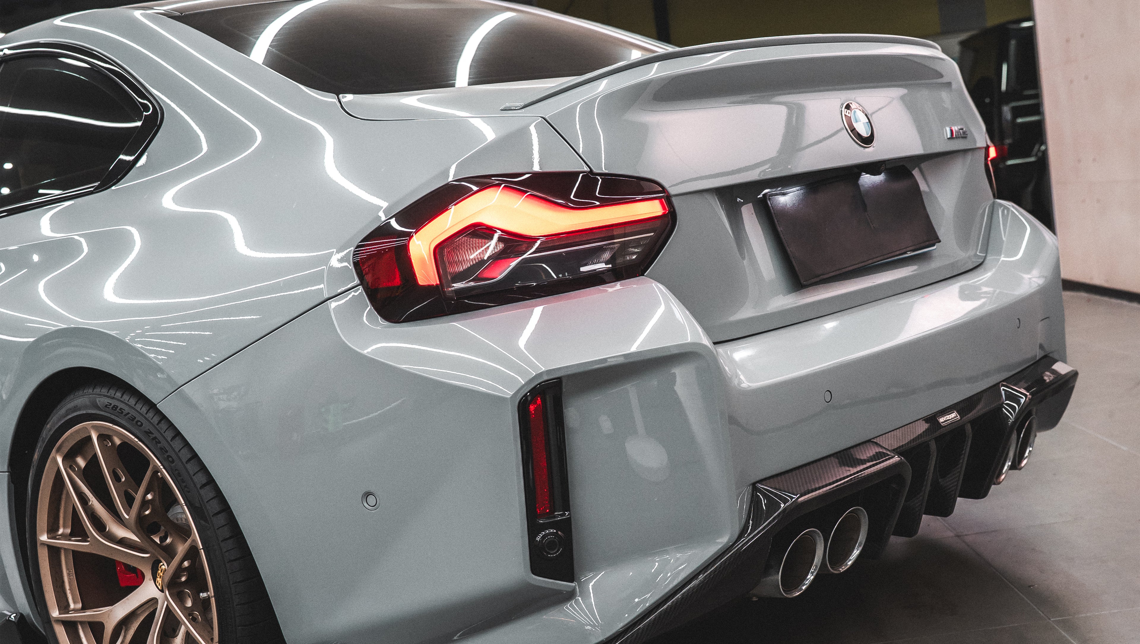 Gray BMW G87 M2 featuring a REVOZPORT carbon fiber front lip, aggressive front aero styling, bronze wheels, and red performance brakes displayed in a modern indoor garage.