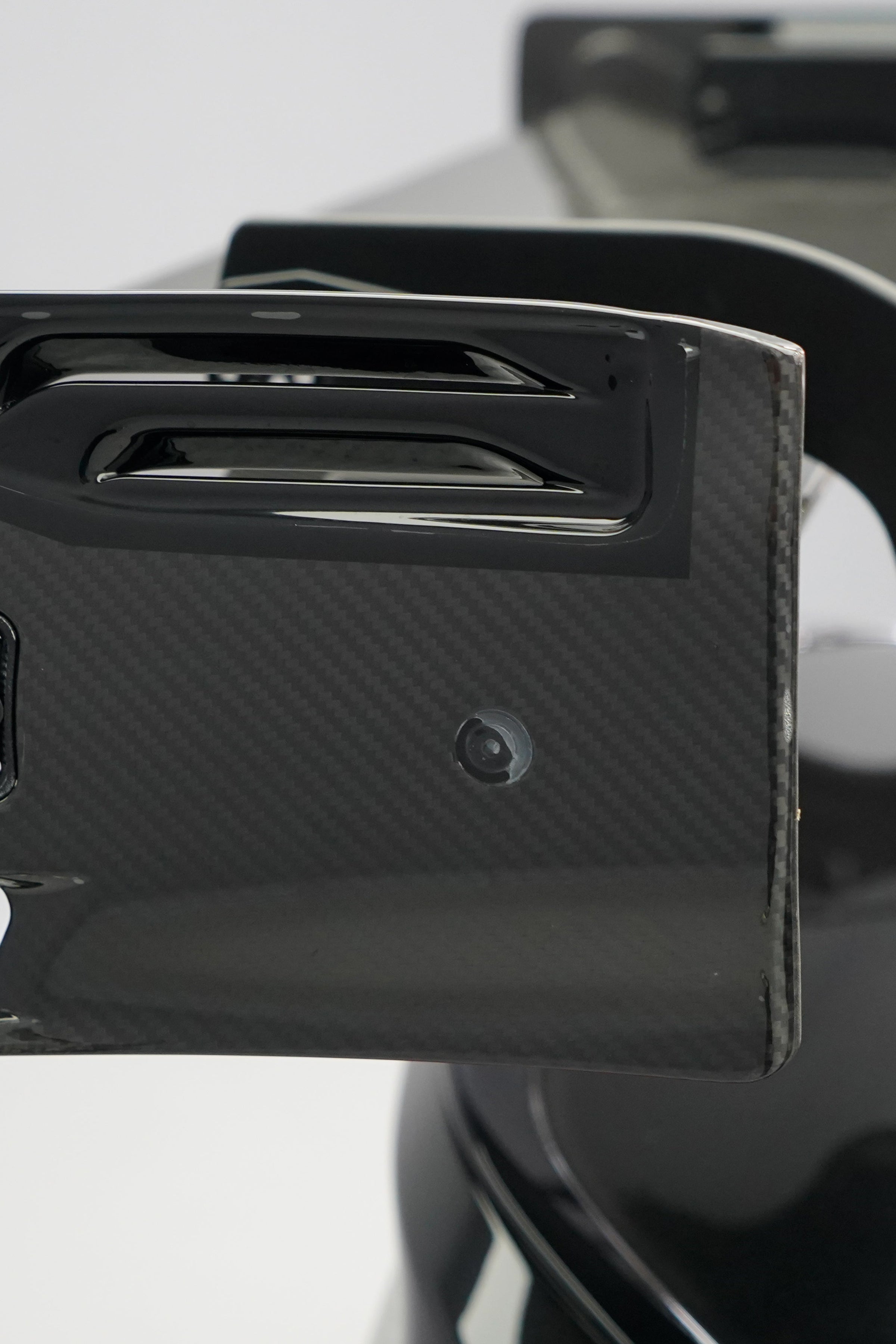 Front close-up of a black BMW G87 M2 featuring an aggressive carbon fiber front bumper, deep front splitter, large air intakes, and gloss carbon fiber aero details in a studio setting.