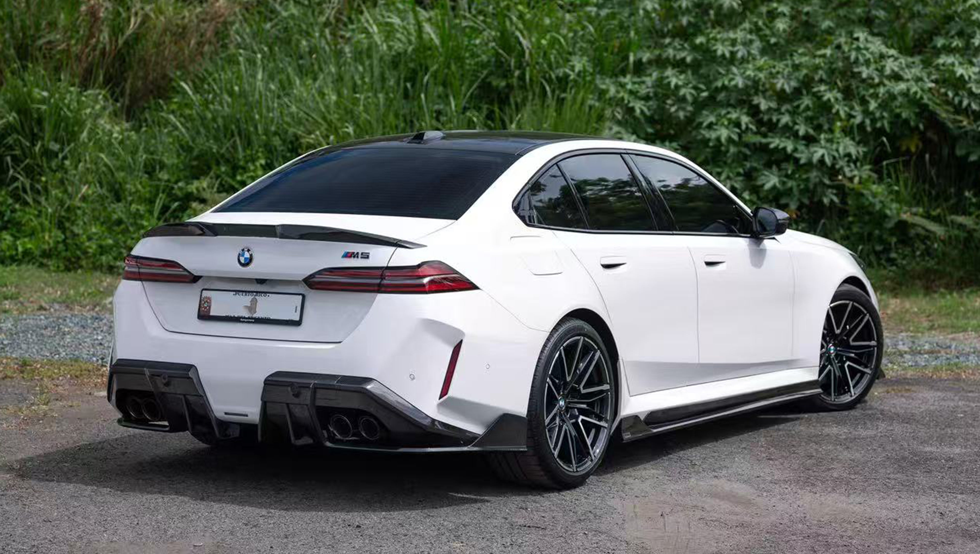 White BMW G90 M5 featuring a REVOZPORT carbon fiber front lip, side skirts, and aggressive front aero styling parked in a natural outdoor setting.