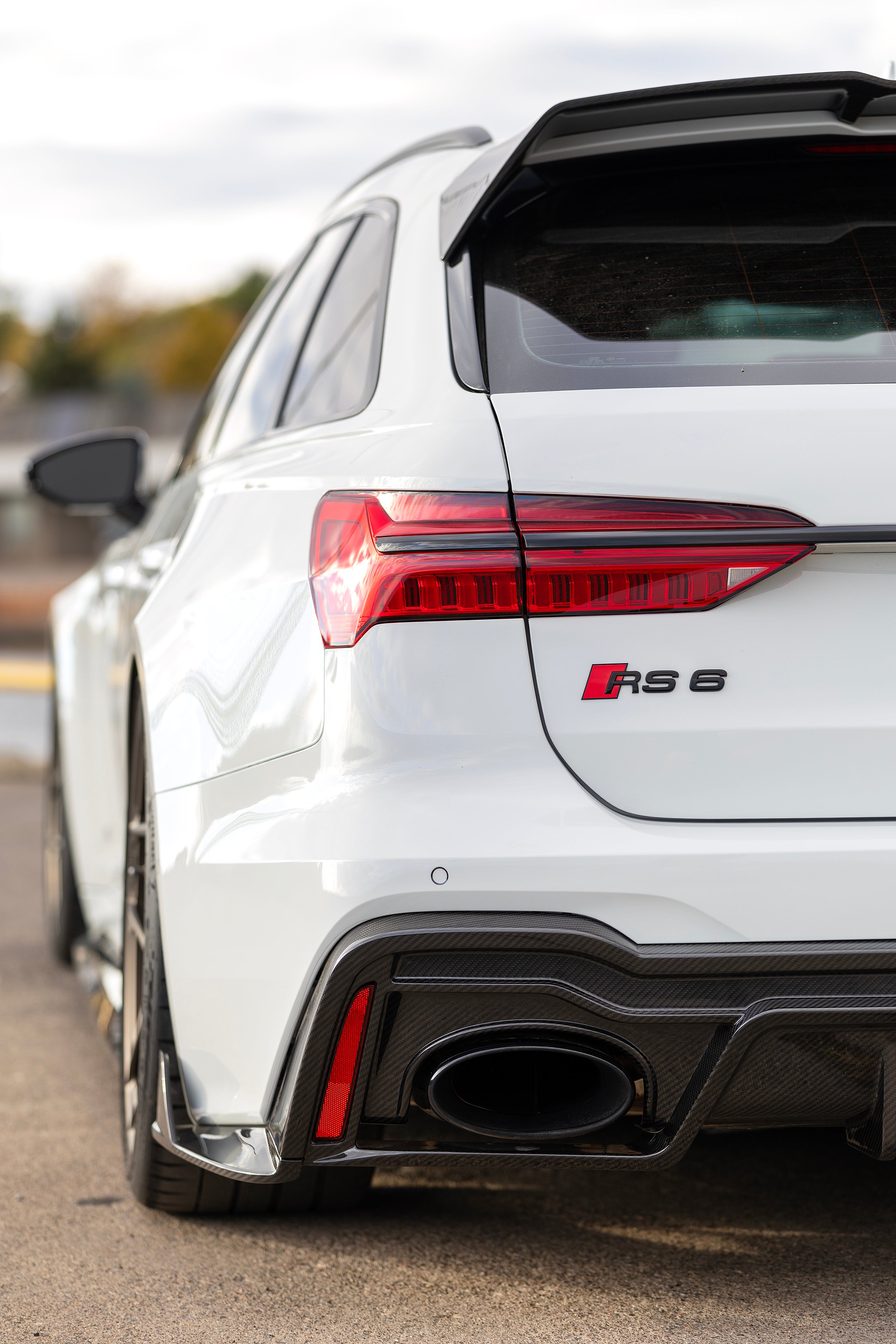 White Audi RS6 Avant featuring a REVOZPORT carbon fiber body kit with front lip, side skirts, and aggressive aero styling parked in front of a historic stone mansion during autumn.