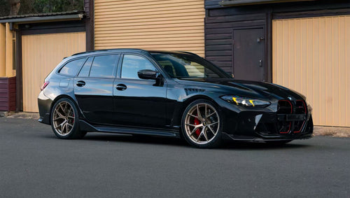Black BMW G81 M3 Touring featuring a REVOZPORT carbon fiber body kit with vented fenders, side skirts, bronze wheels, and red performance brakes parked beside a stone wall.