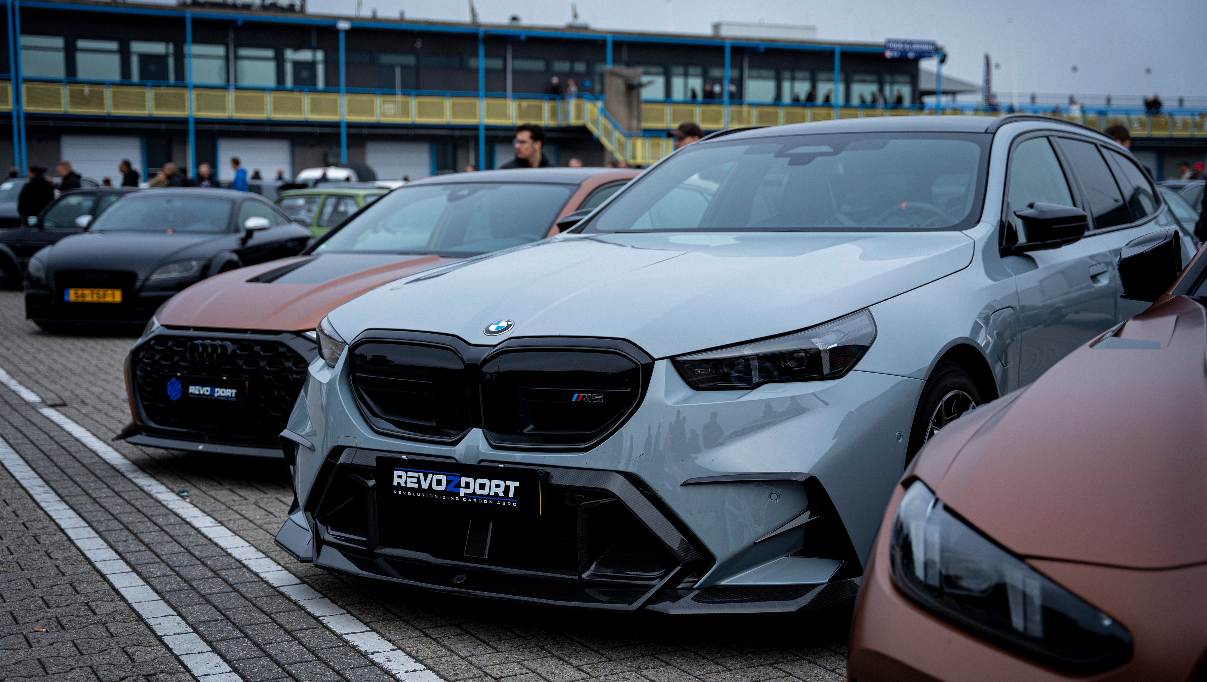 Gray BMW G90 M5 Touring featuring a REVOZPORT carbon fiber front lip, aggressive front aero styling, and black grille displayed at a car event.