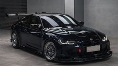 Dark blue BMW G82 M4 equipped with carbon fiber front splitter, side skirts, vented fenders, and a large rear wing, parked inside a modern luxury garage.
