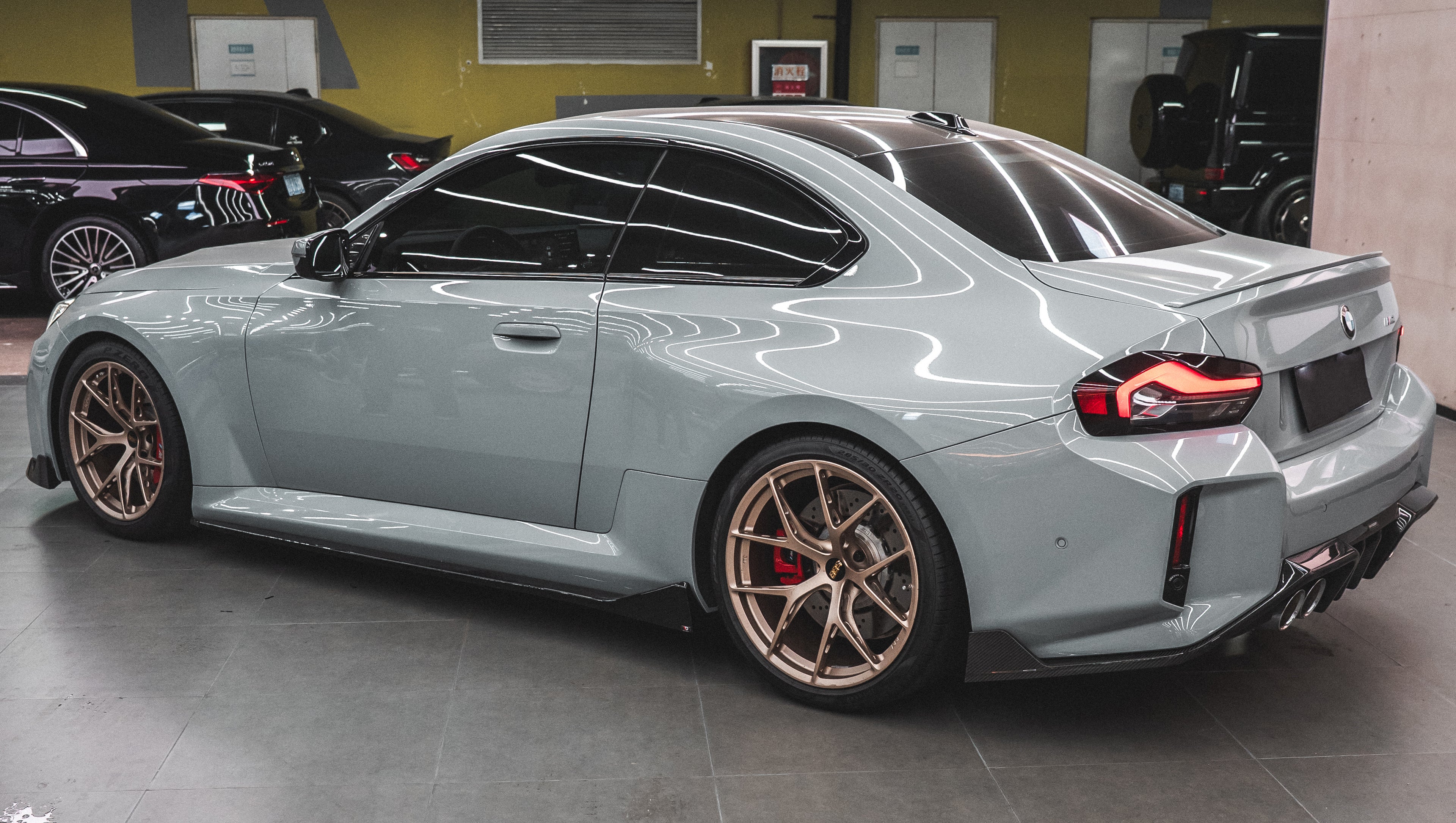 Gray BMW G87 M2 featuring a REVOZPORT carbon fiber front lip, aggressive front aero styling, bronze wheels, and red performance brakes displayed in a modern indoor garage.