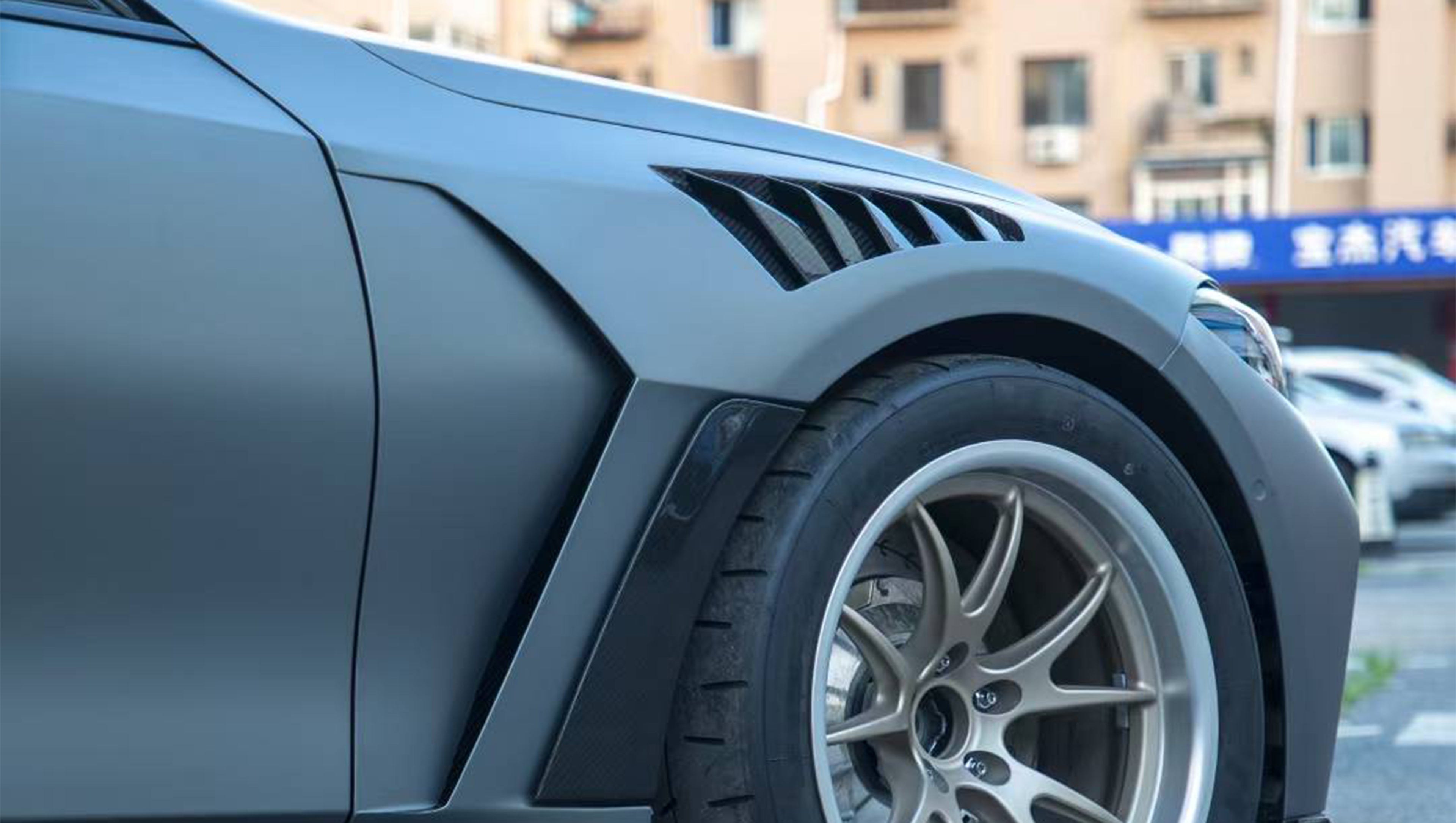 Close-up of a gray BMW G80 M3 featuring a REVOZPORT carbon fiber front lip, vented fenders, side skirts, and aggressive front aero styling in an outdoor parking lot.