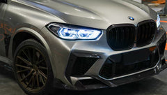 Gray BMW X5M featuring a REVOZPORT carbon fiber body kit with aggressive front lip, enlarged air intakes, side skirts, and sporty aero styling displayed inside a performance workshop.