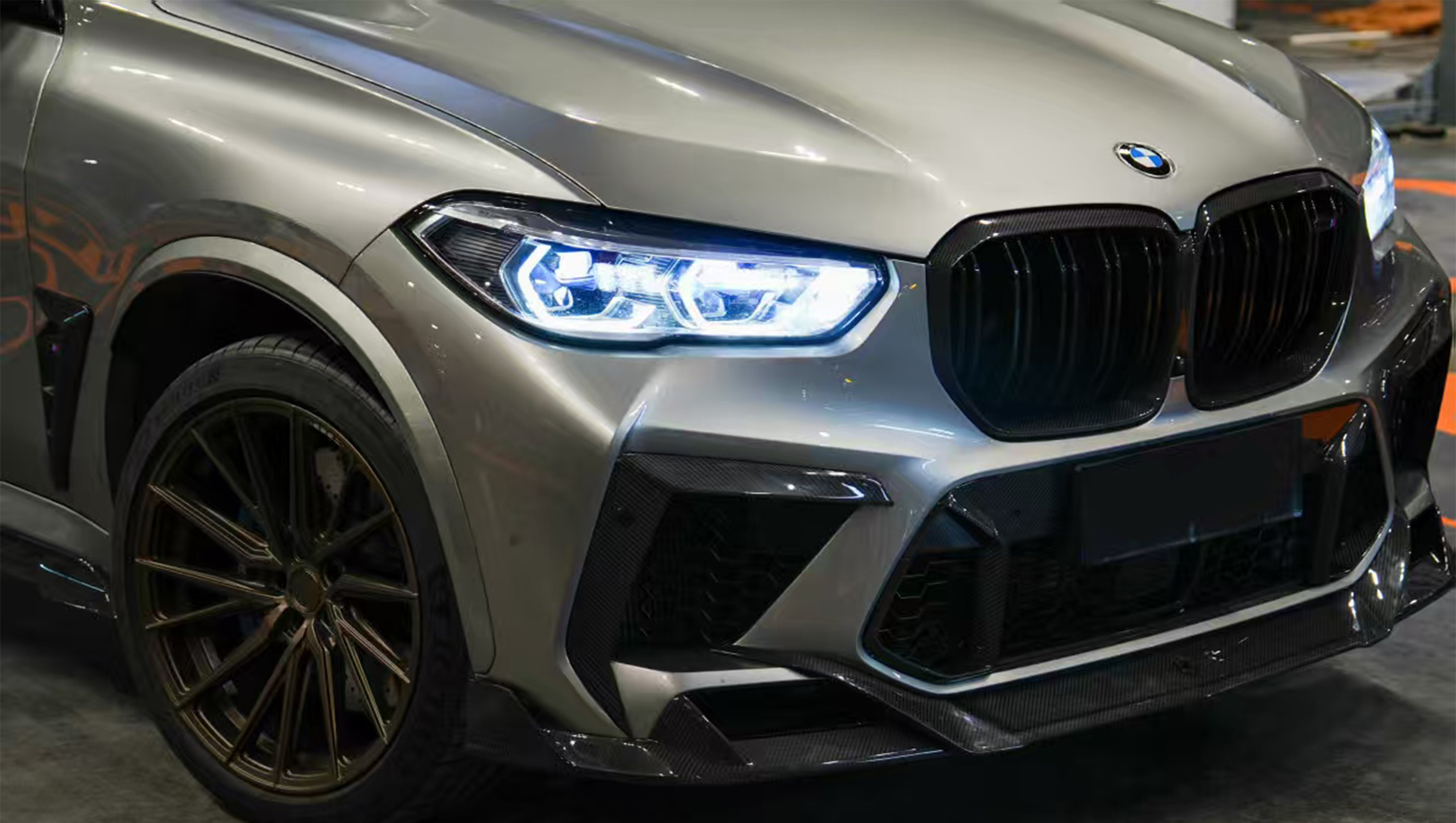 Gray BMW X5M featuring a REVOZPORT carbon fiber body kit with aggressive front lip, enlarged air intakes, side skirts, and sporty aero styling displayed inside a performance workshop.