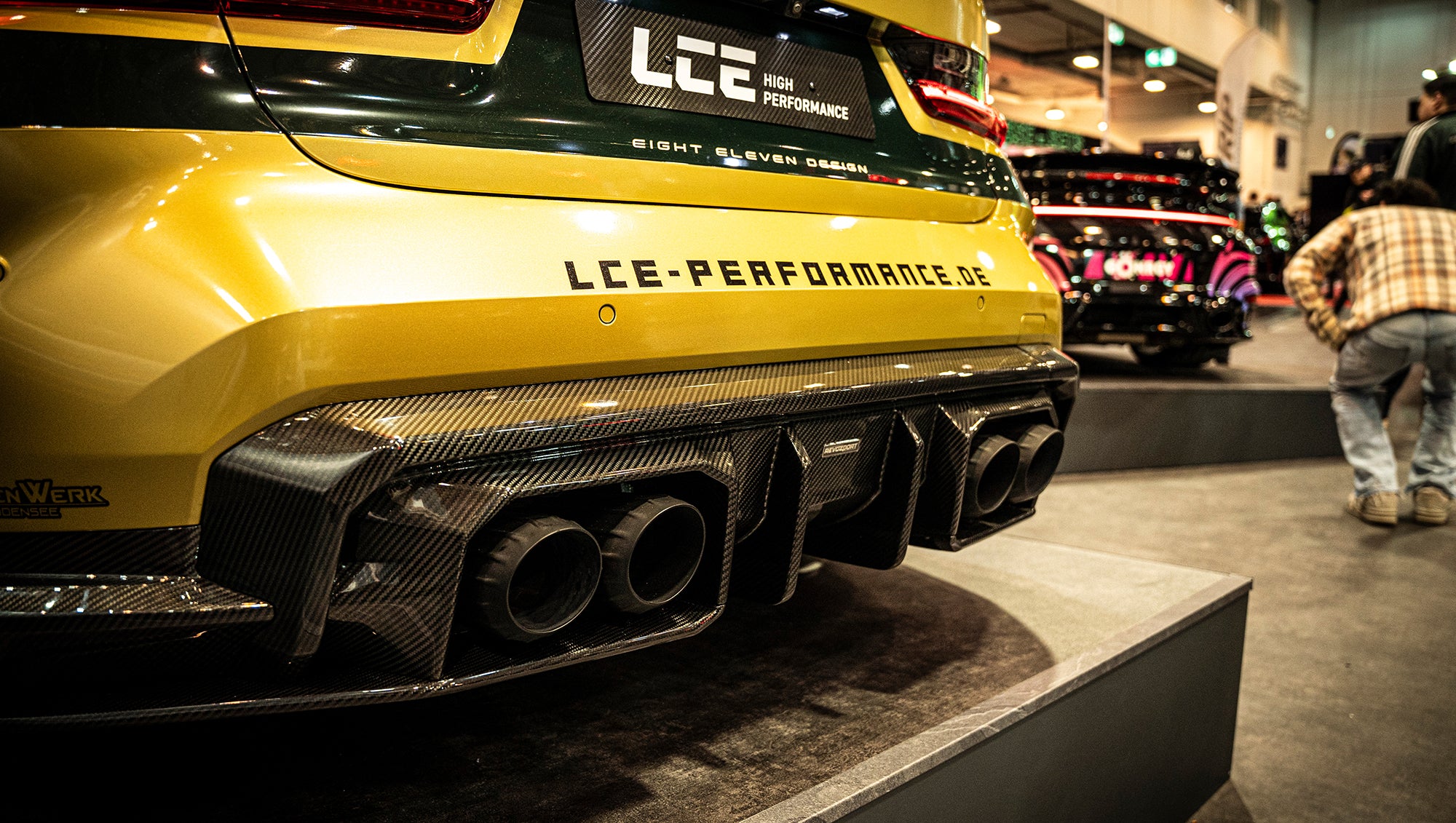 BMW G80 M3 race build with REVOZPORT carbon fiber aero kit, gold wheels, and black-yellow motorsport livery displayed at an indoor automotive exhibition.