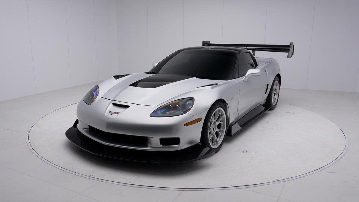 Close-up of a silver wide-body Chevrolet Corvette performance car displayed indoors, featuring aggressive aerodynamic bodywork, carbon fiber front splitter, vented hood, and lightweight multi-spoke wheels. The image highlights the custom motorsport styling and premium carbon fiber aero components under exhibition lighting.