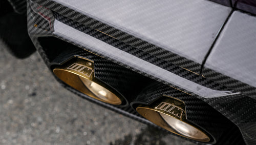Close-up of REVOZPORT carbon fiber rear diffuser with dual gold exhaust tips, highlighting the glossy carbon weave and performance rear aero detail.