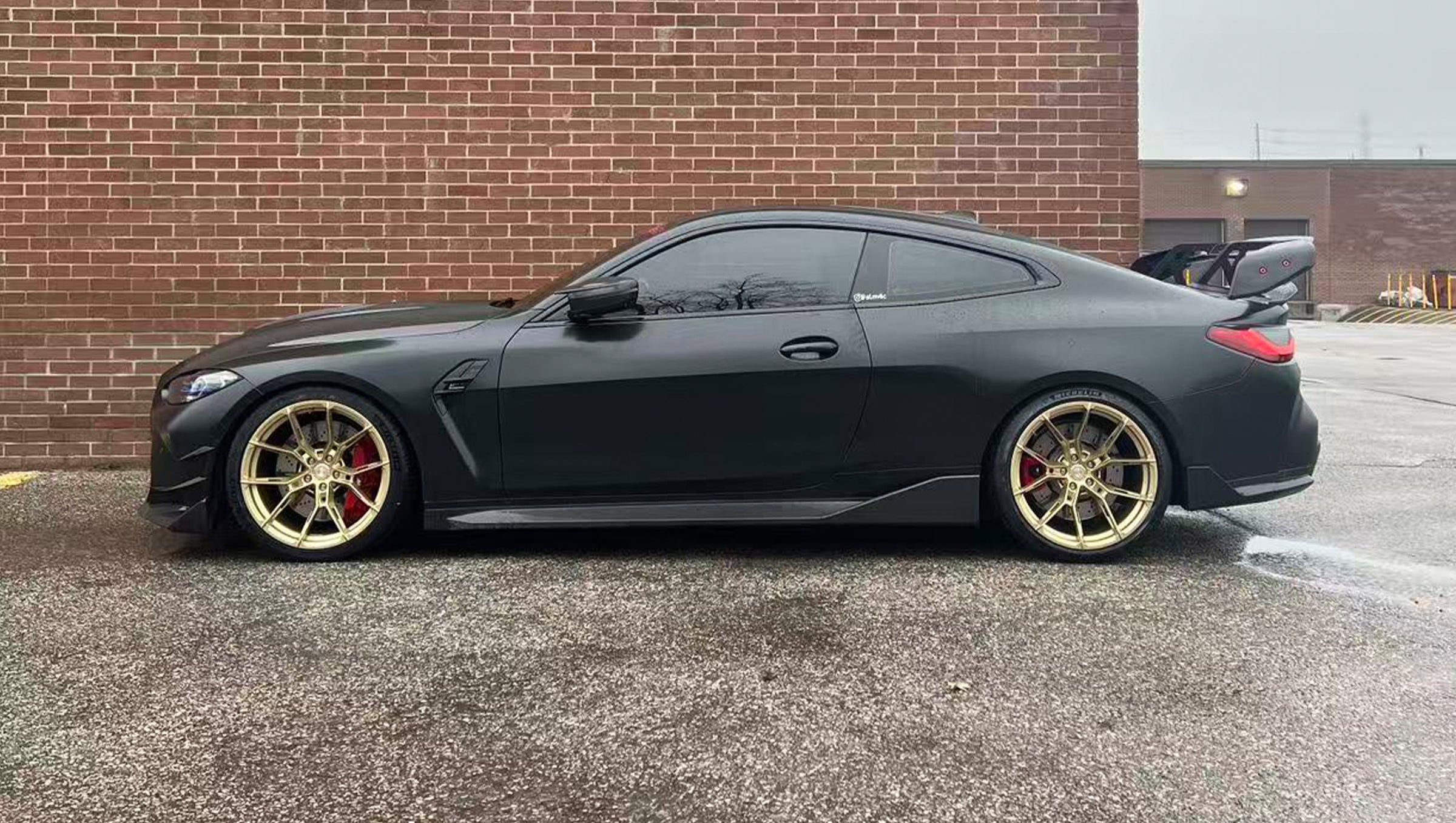 Side view of a matte black BMW G82 M4 featuring REVOZPORT carbon fiber side skirts, front lip, rear wing, and bronze forged wheels parked beside a brick wall.