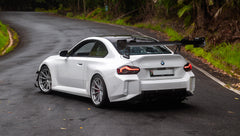 White widebody BMW G82 M4 with aggressive front bumper, canards, deep front splitter, large rear wing, and yellow LED headlights parked on a wet road surrounded by dense tropical forest.