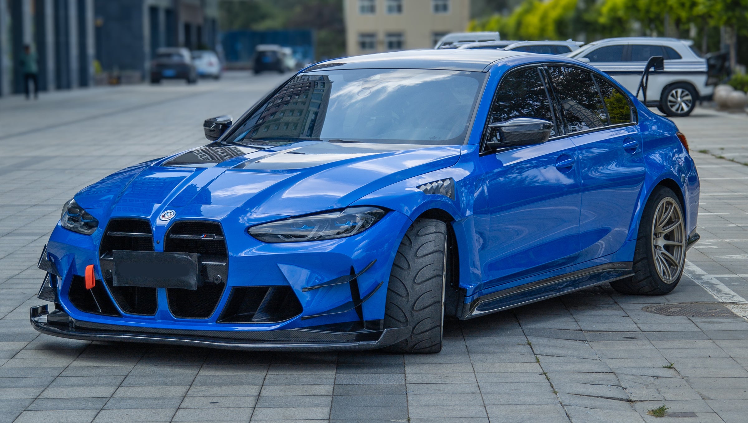 Blue BMW G80 M3 with REVOZPORT carbon fiber front splitter, side skirts, vented fenders, and large rear wing parked in a landscaped outdoor plaza.