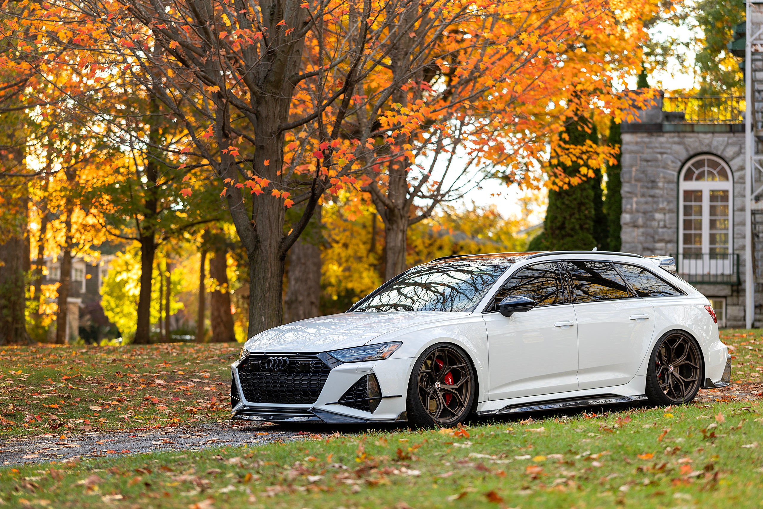 White Audi RS6 Avant featuring a REVOZPORT carbon fiber body kit with front lip, side skirts, and aggressive aero styling parked in front of a historic stone mansion during autumn.