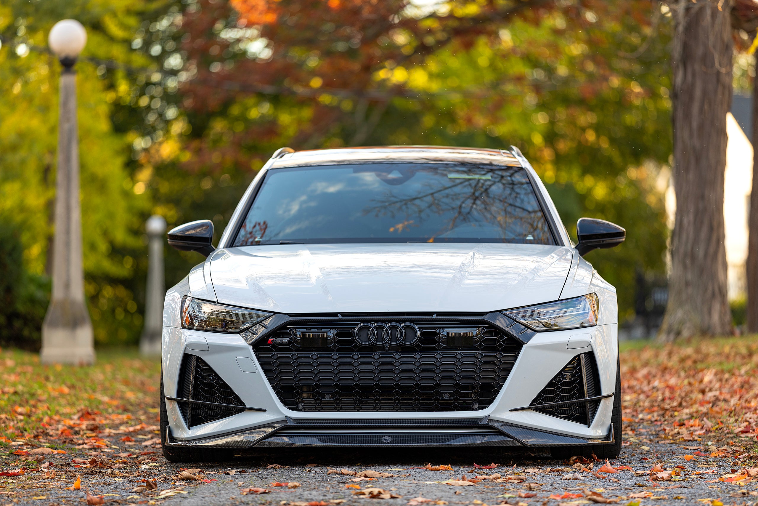 White Audi RS6 Avant featuring a REVOZPORT carbon fiber body kit with front lip, side skirts, and aggressive aero styling parked in front of a historic stone mansion during autumn.