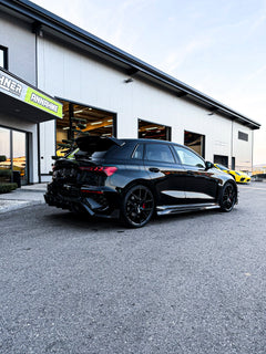 Black Audi RS3 Sportback featuring a REVOZPORT carbon fiber body kit with front lip and side skirts parked on a wet outdoor lot.