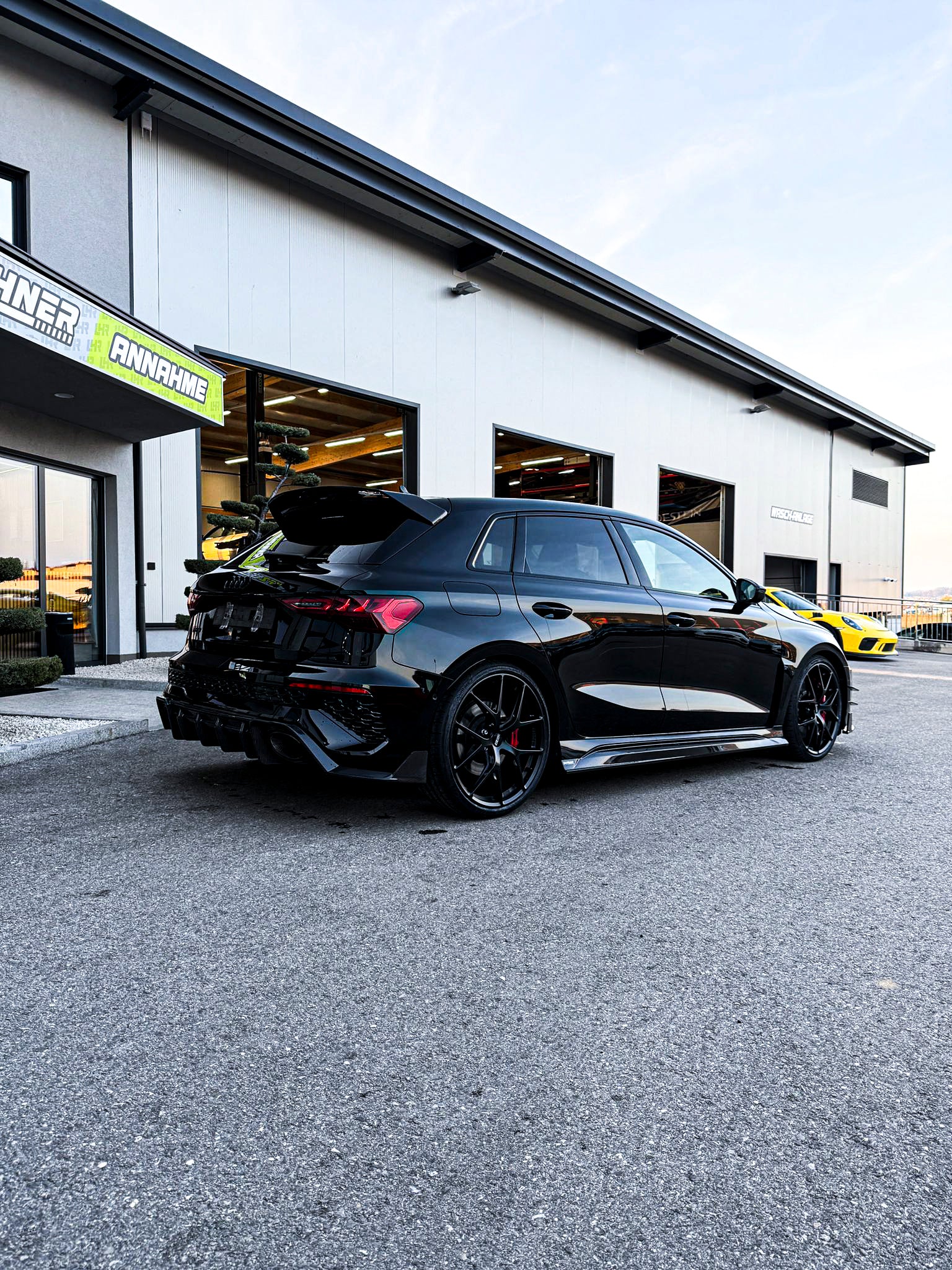Black Audi RS3 Sportback featuring a REVOZPORT carbon fiber body kit with front lip and side skirts parked on a wet outdoor lot.