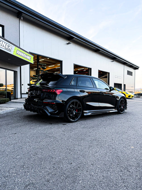 Black Audi RS3 Sportback featuring a REVOZPORT carbon fiber body kit with front lip and side skirts parked on a wet outdoor lot.