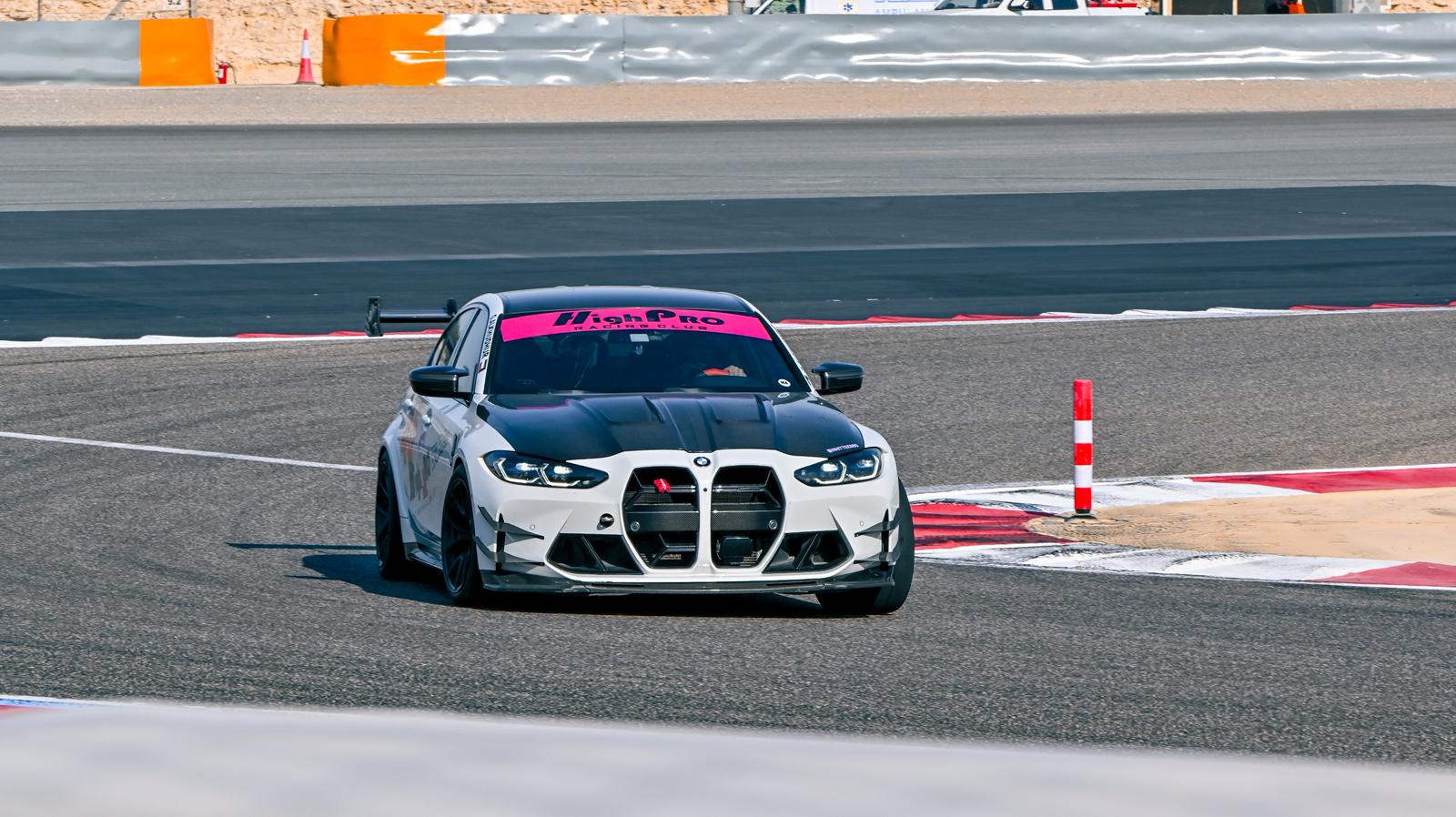 White BMW G80 M3 track car with REVOZPORT carbon fiber aero kit, front canards, rear wing, and racing decals driving on a professional race circuit.